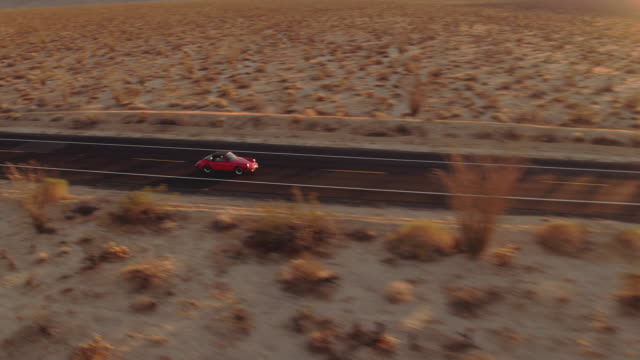 Aerial View Red Classic Car Driving Along Desolate Desert Road At Sunset