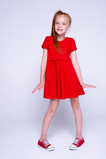 Beautiful little redhead girl in red dress and sneakers posing like model on white background