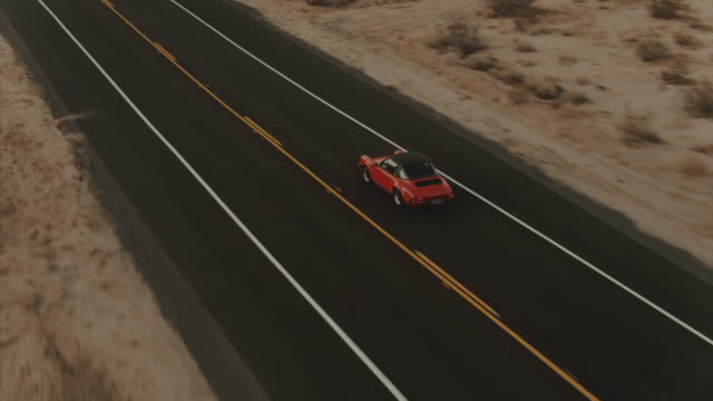 Aerial View Red Classic Car Driving Along Desolate Desert Road At Dusk With Headlights On