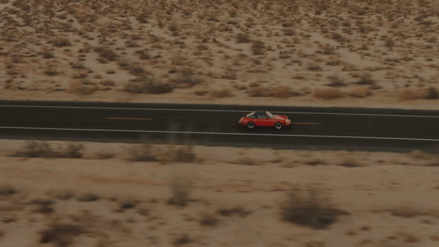 Aerial View Red Classic Car Driving Along Desolate Desert Road At Dusk With Headlights On