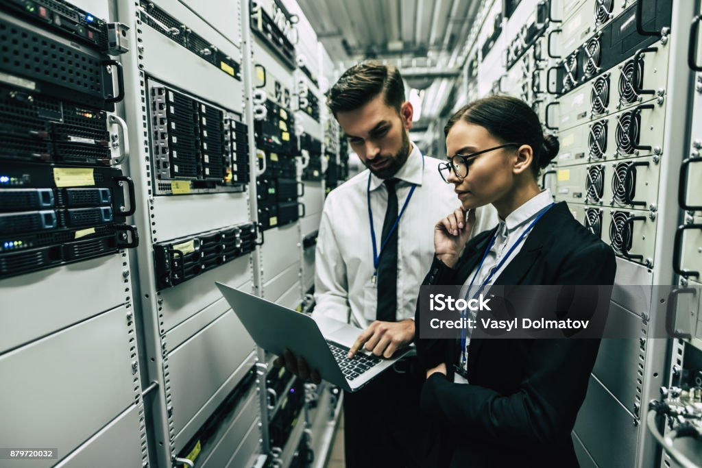 Handsome man and attractive woman are working in data centre. Handsome man and attractive woman are working in data centre with laptop. IT engineer specialists in network server room. Running diagnostics and maintenance. Technicians examining server. Data Center Stock Photo