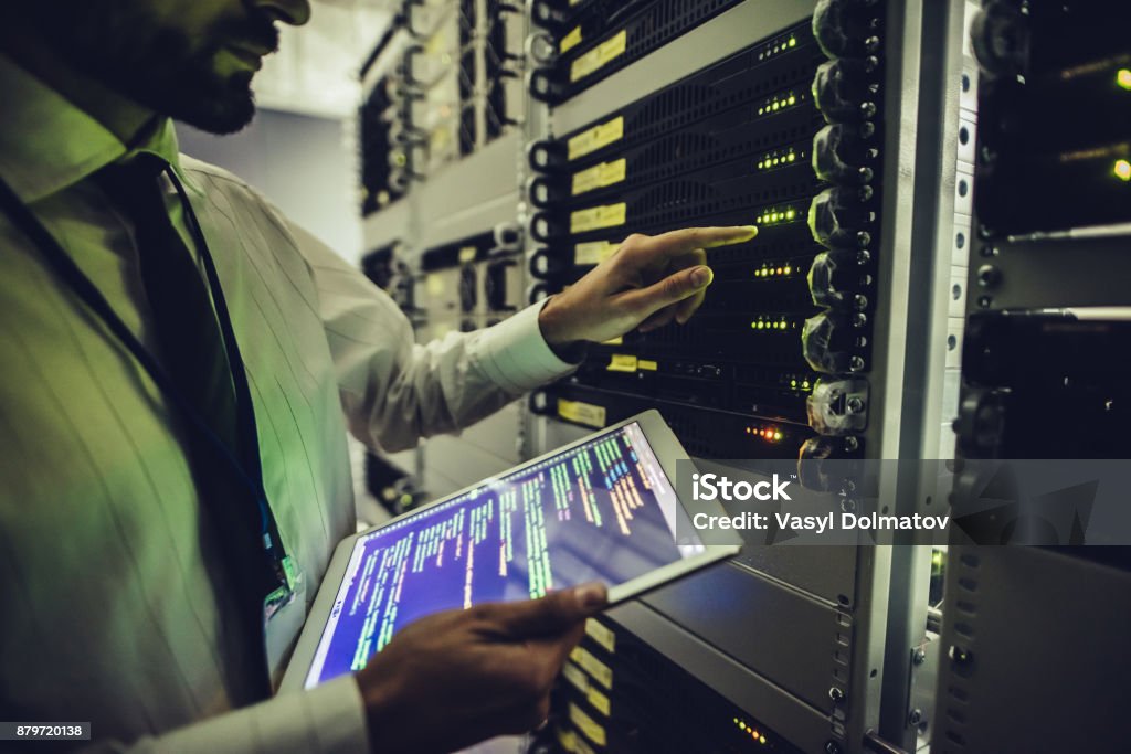 Handsome man is working in data centre with tablet. Handsome man is working in data centre with tablet.IT engineer specialist in network server room.Running diagnostics and maintenance.Technician examining server in big data center full of rack servers Network Server Stock Photo