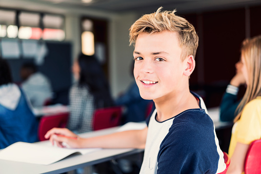 Portrait of blond teenage boy sitting with friends. Happy male student is in classroom. He is looking over shoulder at high school.