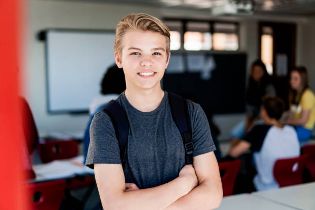 Smiling blond teenage boy with arms crossed Portrait of smiling teenage boy standing against friends. Happy male blond student is with arms crossed in classroom. He is wearing casual clothing in school building. 15 year old blonde girl stock pictures, royalty-free photos & images