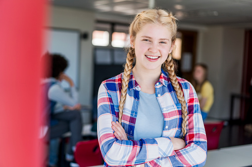 Portrait of smiling blond teenager with arms crossed. Happy female student is with long braided hair standing against friends. She is in classroom of high school building.