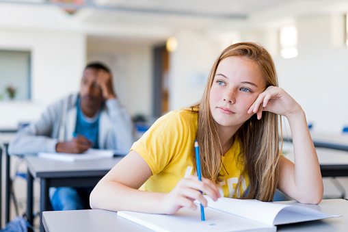 Thoughtful teenage girl leaning on desk. Female student is sitting with pencil and book in classroom. She is against male friend at school.