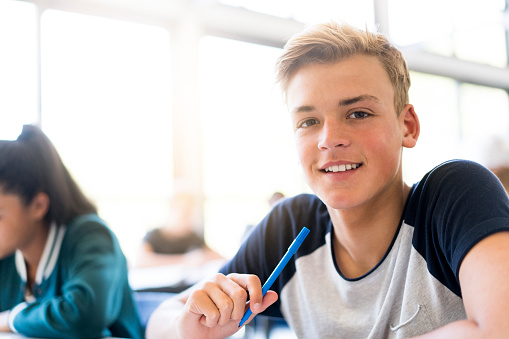 Portrait of smiling blond male student in classroom. Happy teenage boy is sitting by female friend. He is wearing casual at school.