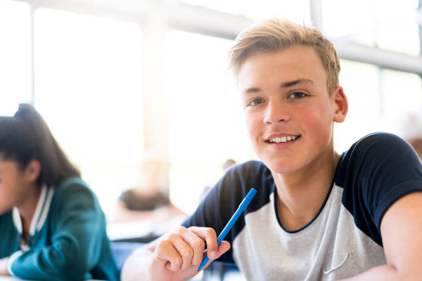 Smiling male teenage student sitting in classroom Portrait of smiling blond male student in classroom. Happy teenage boy is sitting by female friend. He is wearing casual at school. 15 year old blonde girl stock pictures, royalty-free photos & images