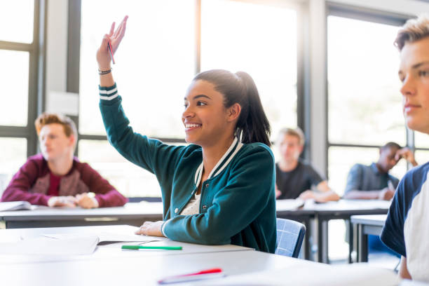 Smiling teenage student raising hand in classroom Smiling teenage girl raising hand in classroom. Happy female student is sitting amidst male friends. They are wearing casuals at school. hand raised classroom student high school student stock pictures, royalty-free photos & images