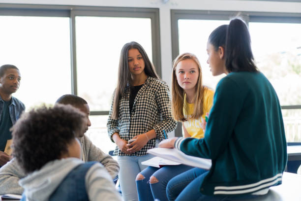 Multi-ethnic students discussing in classroom High school students are discussing against window. Multi-ethnic male and females are in classroom. They are wearing casuals at school. 16 17 years photos stock pictures, royalty-free photos & images