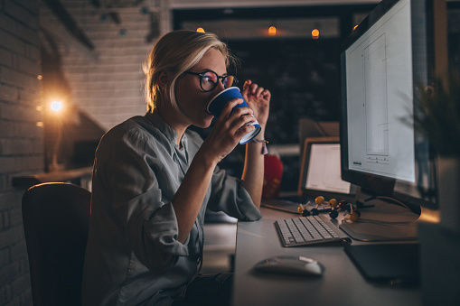 Photo of a young woman sitting at her desktop computer, having some hot coffee to help her trough a late night shift