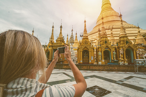 Young woman traveling in Myanmar takes a picture of the Shwedagon pagoda with a mobile phone. People travel youth culture concept