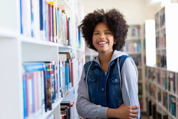 Smiling teenage female student in library Portrait of cheerful teenage girl standing by bookshelf. Smiling female student is with afro hairstyle in library. She is wearing casuals at high school. happy young teens stock pictures, royalty-free photos & images