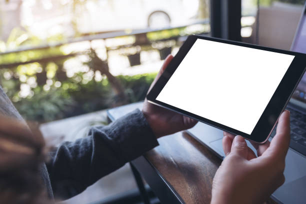 immagine mockup delle mani che tengono il tablet pc nero con schermo bianco bianco e laptop su sfondo tavolo di legno nel caffè - human hand computer equipment household equipment foto e immagini stock