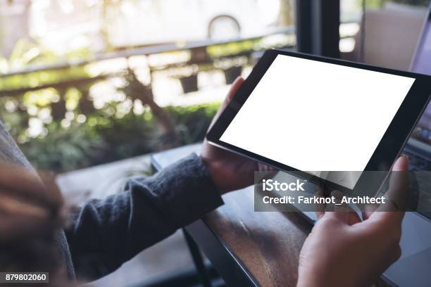 Mockup Image Of Hands Holding Black Tablet Pc With White Blank Screen And Laptop On Wooden Table Background In Cafe Stock Photo - Download Image Now