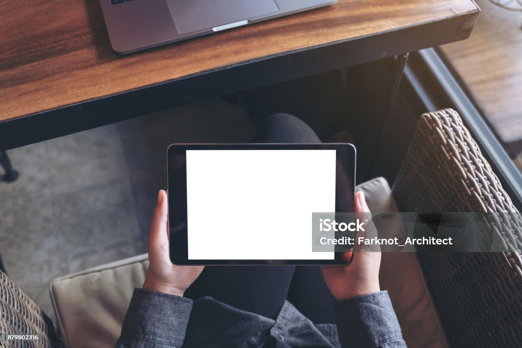 Mockup image of woman's hands holding black tablet pc with blank white screen on thigh with laptop in modern cafe Digital Tablet Stock Photo