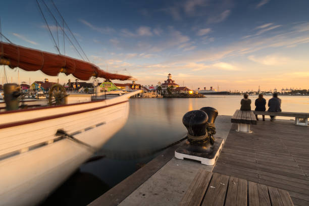 Tourists enjoy a beautiful sunset as they sit on a bench on the end of Pine Avenue Pier in Rainbow Harbor in the heart of downtown Long Beach, CA. They are enjoying the view of Shoreline Village and the Queen Mary in the distance. Shot with a long exposore technique.