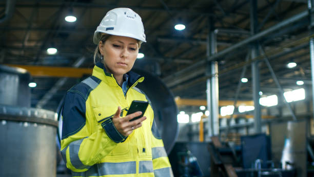 Female Industrial Worker in the Hard Hat Uses Mobile Phone While Walking Through Heavy Industry Manufacturing Factory. In the Background Various Metalwork Project Parts Lying Female Industrial Worker in the Hard Hat Uses Mobile Phone While Walking Through Heavy Industry Manufacturing Factory. In the Background Various Metalwork Project Parts Lying mobile phone finance business technology stock pictures, royalty-free photos & images