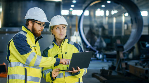 istock Male and Female Industrial Engineers in Hard Hats Discuss New Project while Using Laptop. They Make Showing Gestures.They Work in a Heavy Industry Manufacturing Factory. 879813802