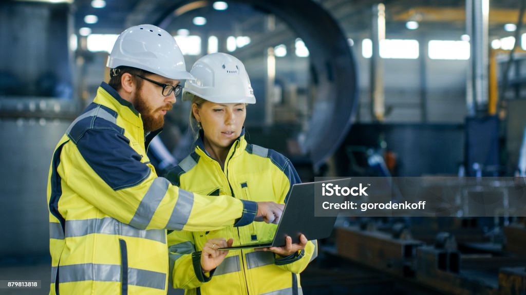 Male and Female Industrial Engineers in Hard Hats Discuss New Project while Using Laptop. They Make Showing Gestures.They Work in a Heavy Industry Manufacturing Factory. Engineer Stock Photo