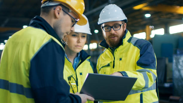 istock Three Industrial Engineers Talk with Factory Worker while Using Laptop. They Work at the Heavy Industry Manufacturing Facility. 879813904