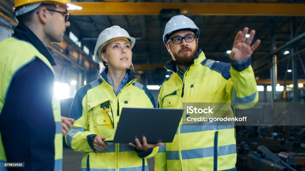 Male and Female Industrial Engineers Talk with Factory Worker while Using Laptop. They Work at the Heavy Industry Manufacturing Facility. Safety Stock Photo