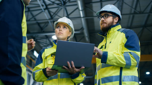 istock Male and Female Industrial Engineers Talk with Factory Worker while Using Laptop. They Work at the Heavy Industry Manufacturing Facility. Low Angle Shot. 879813936