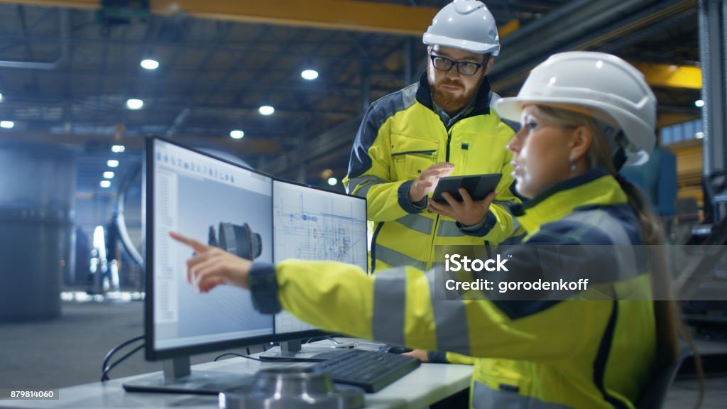 Dentro de la fábrica de industria pesada mujer Ingeniero Industrial trabaja en ordenador Personal ella diseños 3D turbina modelo, su colega masculino habla con ella y utiliza la computadora de la tableta. - Foto de stock de Ingeniero libre de derechos