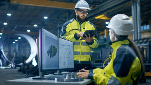 istock Inside the Heavy Industry Factory Female Industrial Engineer Works on Personal Computer She Designs 3D Turbine Model, Her Male Colleague Talks with Her and Uses Tablet Computer. 879814062