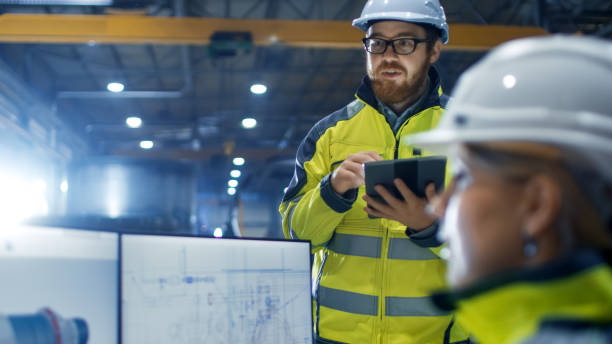 istock Inside the Heavy Industry Factory Female Industrial Engineer Works on Personal Computer She Designs 3D Engine Model, Her Male Colleague Talks with Her and Uses Tablet Computer. 879814074