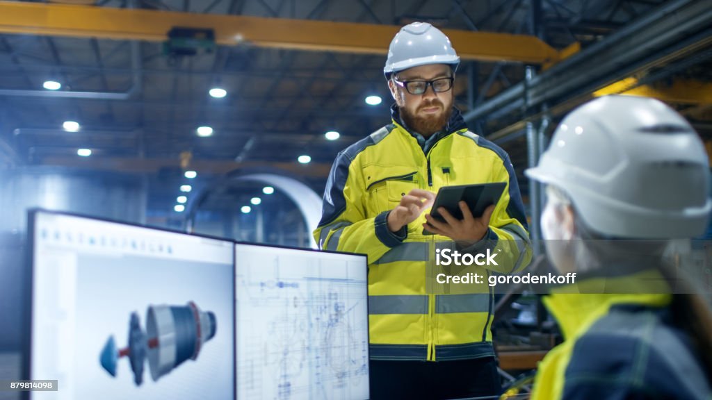 Inside the Heavy Industry Factory Female Industrial Engineer Works on Personal Computer She Designs 3D Engine Model, Her Male Colleague Talks with Her and Uses Tablet Computer. Engineer Stock Photo