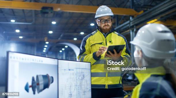 Inside The Heavy Industry Factory Female Industrial Engineer Works On Personal Computer She Designs 3d Engine Model Her Male Colleague Talks With Her And Uses Tablet Computer Stock Photo - Download Image Now