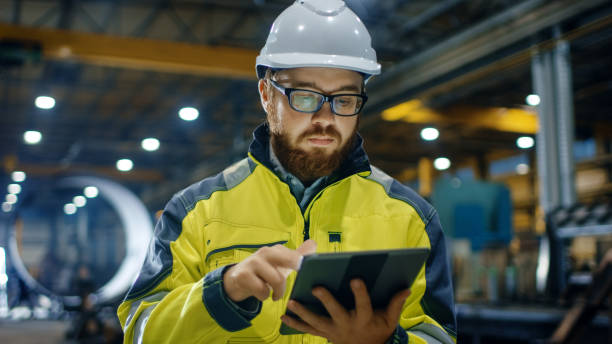 engenheiro industrial no capacete, jaqueta de segurança usa computador tablet touchscreen. ele trabalha na indústria pesada, fabricação de fábrica. - gear equipment industry factory - fotografias e filmes do acervo