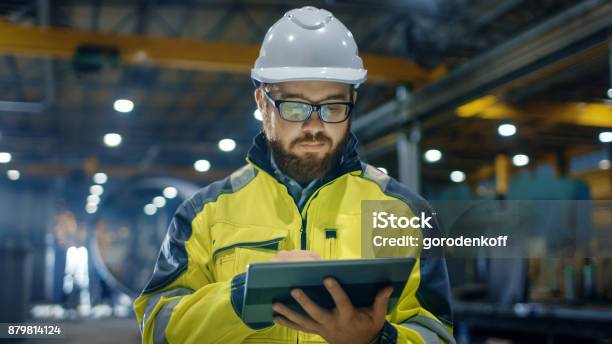 Industrial Engineer In Hard Hat Wearing Safety Jacket Uses Touchscreen Tablet Computer He Works At The Heavy Industry Manufacturing Factory Stock Photo - Download Image Now