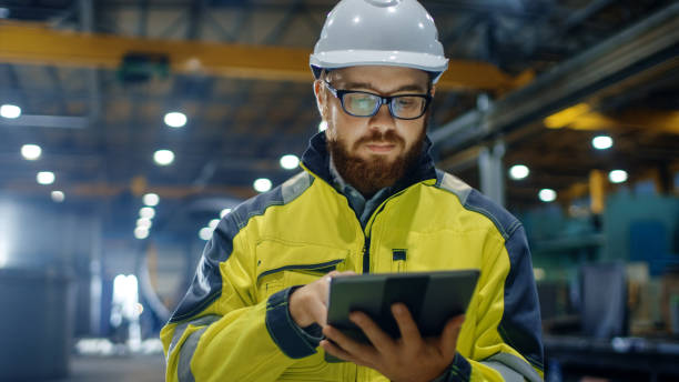 istock Industrial Engineer in Hard Hat Wearing Safety Jacket Uses Touchscreen Tablet Computer. He Works at the Heavy Industry Manufacturing Factory. 879814136
