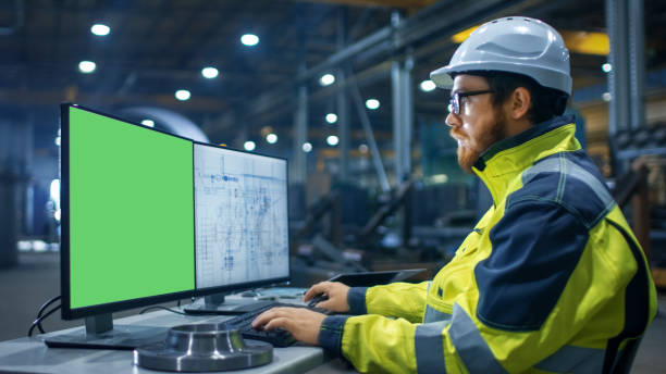 istock Inside the Heavy Industry Factory Industrial Engineer Works on the Personal Computer with Two Screen. One is Green Mock-up Screen and Other with Blueprints. 879814198