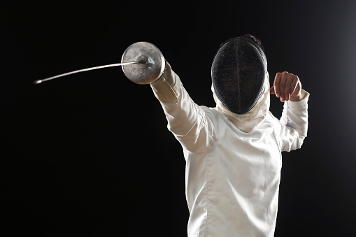 Unrecognizable young man in fencing equipment - uniform, face mask and foil sword on black background.