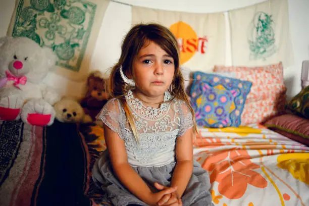 Adorable little girl sits on her bed in a colorful childhood bedroom looking sad and forlorn