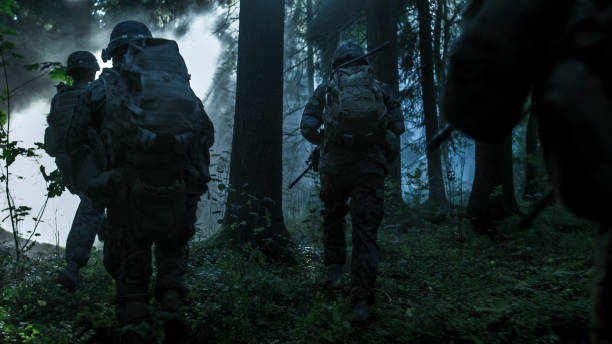 istock Fully Equipped Soldiers Wearing Camouflage Uniform Attacking Enemy, Rifles Ready to Shoot. Military Operation in Action, Squad Running in Formation Through Dark Dense Smokey Forest. 879913020