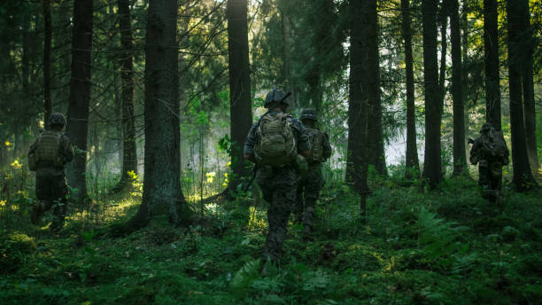 istock Fully Equipped Soldiers Wearing Camouflage Uniform Attacking Enemy, Rifles Ready to Shoot. Military Operation in Action, Squad Running in Formation Through Dense Smokey Forest. Back View Footage. 879913080