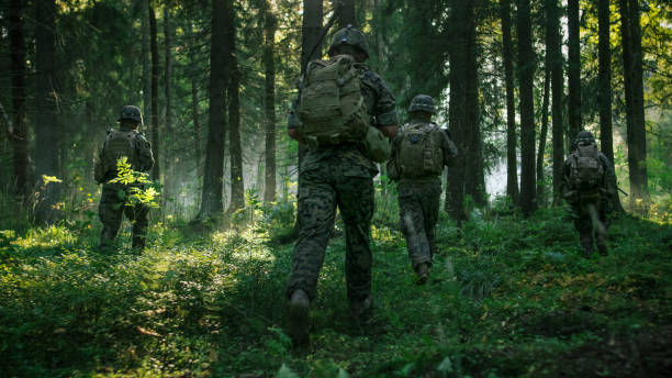 istock Fully Equipped Soldiers Wearing Camouflage Uniform Attacking Enemy, Rifles Ready to Shoot. Military Operation in Action, Squad Running in Formation Through Dense Smokey Forest. Back View Footage. 879913092