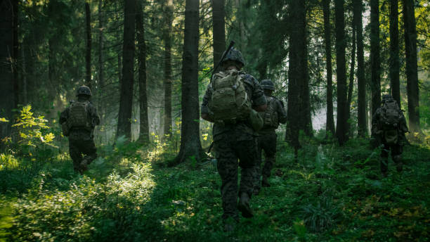 istock Fully Equipped Soldiers Wearing Camouflage Uniform Attacking Enemy, Rifles Ready to Shoot. Military Operation in Action, Squad Running in Formation Through Dense Smokey Forest. Back View Footage. 879913108