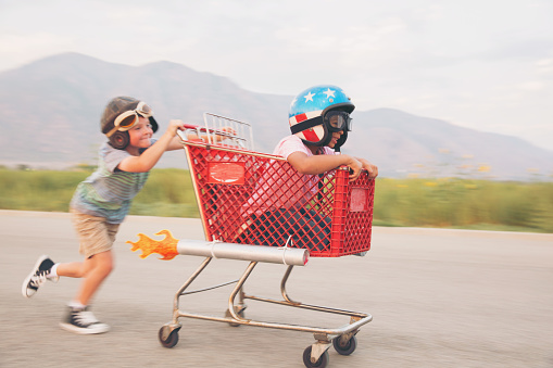 Two young boys wearing racing helmets and racing goggles stand on a rural street for some racing. One is sitting in a shopping cart with rockets strapped on the side. They are looking at the camera excited for the upcoming race. Image taken in Utah, USA.