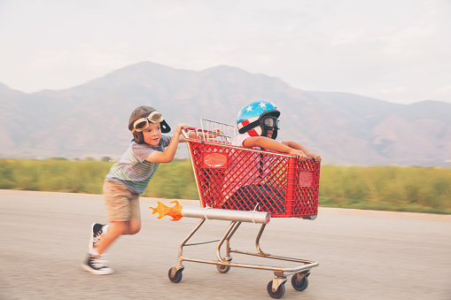 Two young boys wearing racing helmets and racing goggles stand on a rural street for some racing. One is sitting in a shopping cart with rockets strapped on the side. They are looking at the camera excited for the upcoming race. Image taken in Utah, USA.