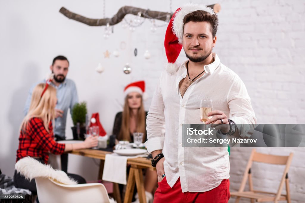 Shot of happy friends enjoying holidays. Focus on the man in the foreground in a red Christmas hat Shot of happy friends enjoying holidays. Focus on the man in the foreground in a red Christmas hat. Man with a glass of champagne in a white room 2015 Stock Photo