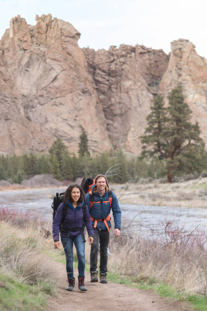 Two longtime friends go backpacking in the wilderness together. She is in front. They are both smiling. There is a small river and a large rock formation rising up behind them.