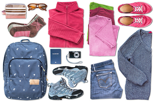 A view from above of unisex travel essentials such as a backpack, shoes, clothes, a digital camera, sunglasses, cosmetic bag and a passport, organized to fill all the space on the white background