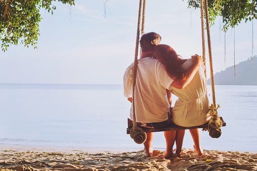 romantic holidays for two, affectionate couple sitting together on the beach on swing, silhouette of man hugging woman