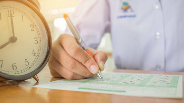 istock Students taking exams writing on optical form of standardized exam near Alarm clock with  holding yellow pen for final examination in secondary school, college university classroom, Education concept 880185008