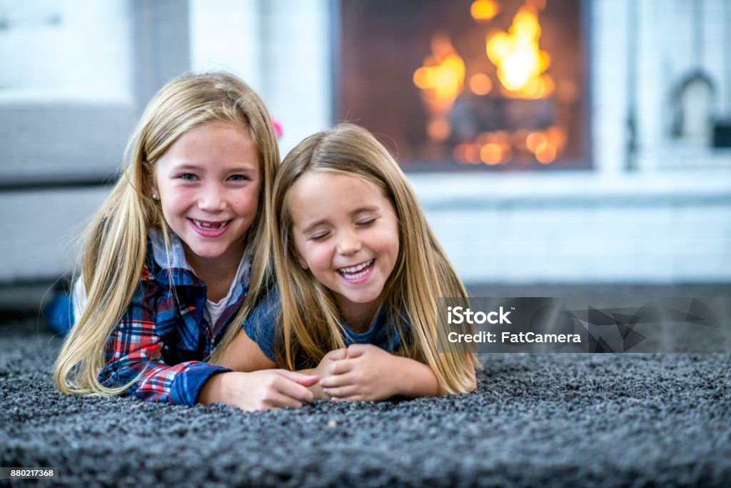 Two Sisters Lying On The Floor Two Caucasian sisters are lying on the carpet in their living room. One girl is laughing and the other is smiling at the camera. There is a fireplace in the background. Carpet - Decor Stock Photo